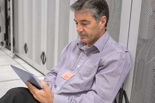 An IT consultant sitting in a server room using a tablet to analyze cybersecurity measures.